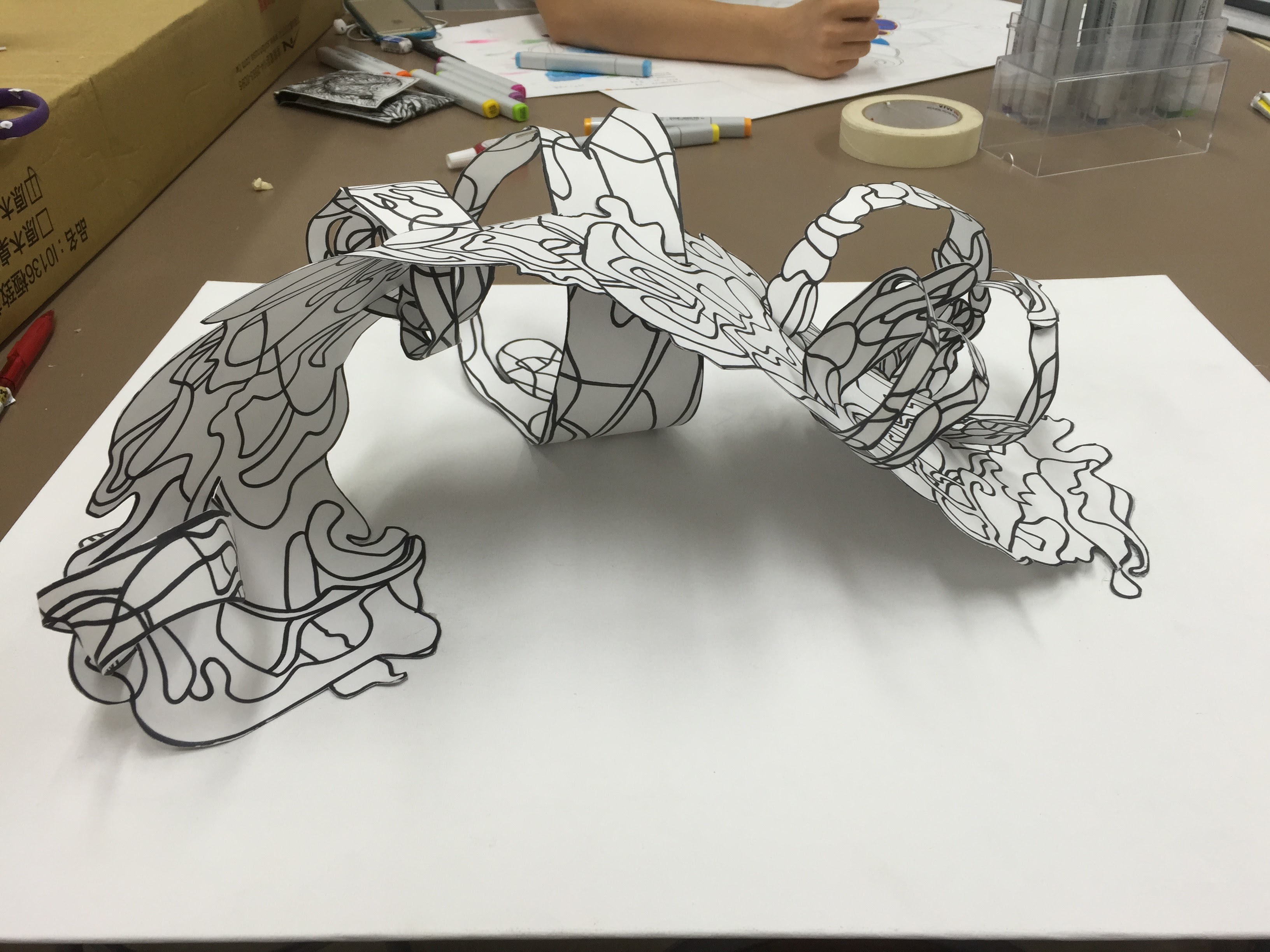 Angie’s paper sculpture: a swirling, ribbon-like 3D form on a desk with art supplies in the background.
